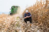 A farmer examines an ear of corn with a farming machine behind him.