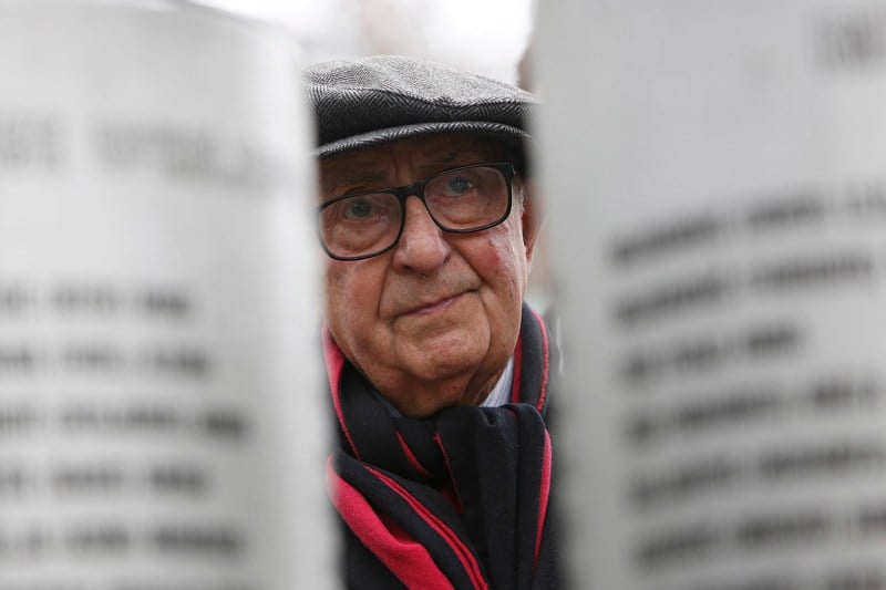 Theodor Meron, an elderly man wearing a hat and glasses, is framed between two columns with names etched into them as he stands at a memorial for children killed in Sarajevo.
