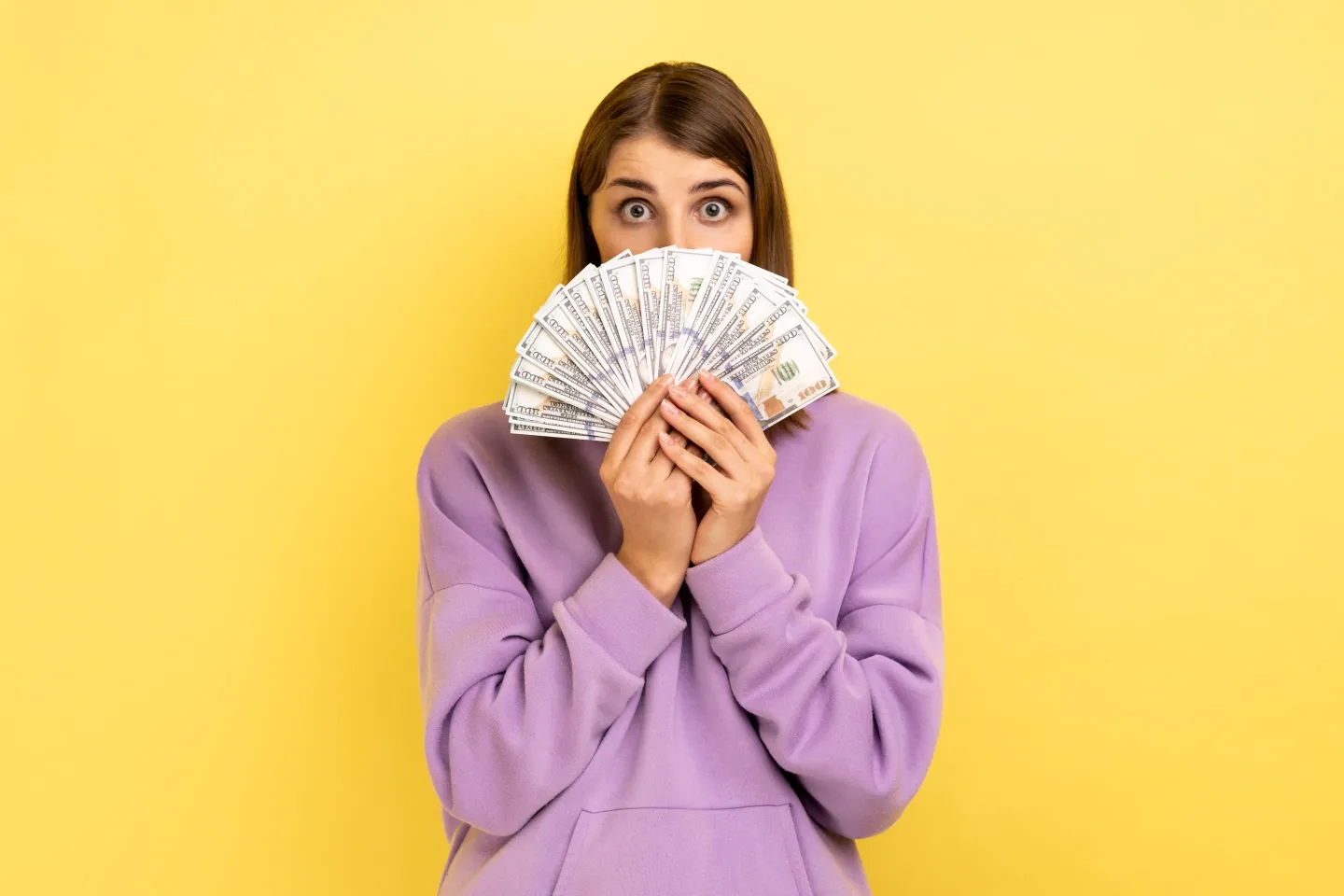 Portrait of amazed woman covering half of face with money and peeking out of dollars, excited to hold lot of cash, wearing purple hoodie. Indoor studio shot isolated on yellow background.