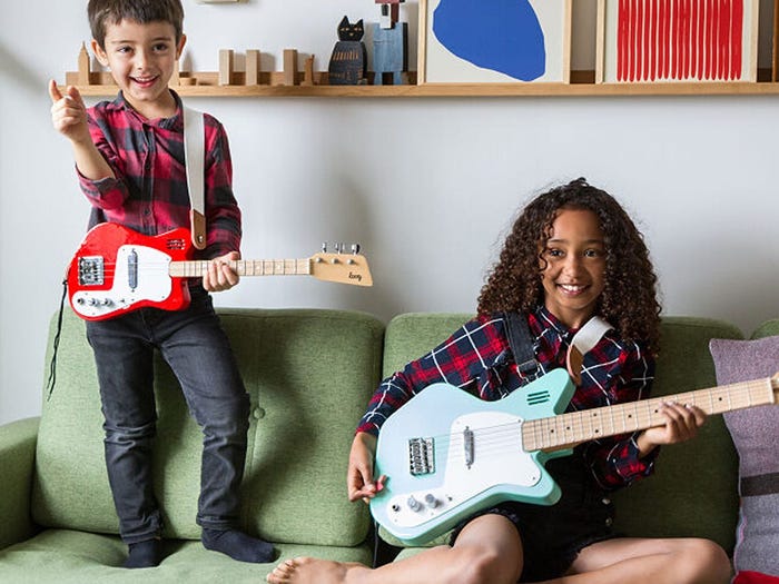 A boy standing and a girl sitting on a couch both holding guitars
