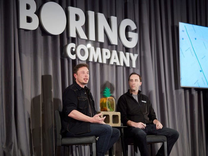 Two men sit on a stage in front of a gray curtain with The Boring Company logo.