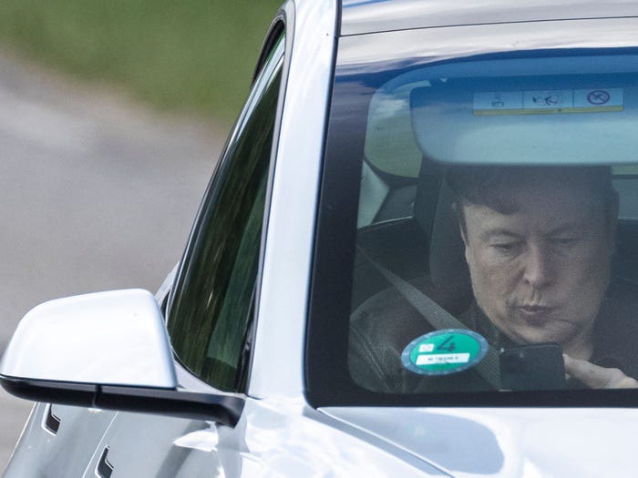 Elon Musk sits inside a white car looking at a cell phone