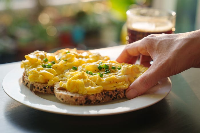 A cropped image of a woman's hand holding a piece of toasted bread with scrambled eggs on top, as she enjoys a healthy breakfast in the morning - stock photo