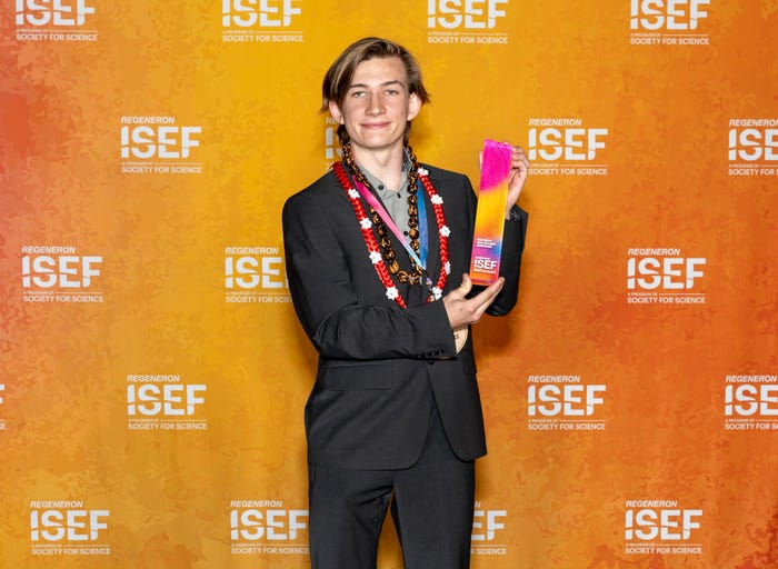 smiling young man wearing a suit and hawaiian style necklaces hold a long rectangular pink and yellow trophy in front of an orange wall with Regeneron ISEF written on it