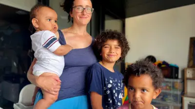 A woman with glasses and curly brown hair, Thea, and her three children: Isaac, a baby on her hip, Moses, aged 9 and smiling broadly, and JJ, aged two, closest to the camera