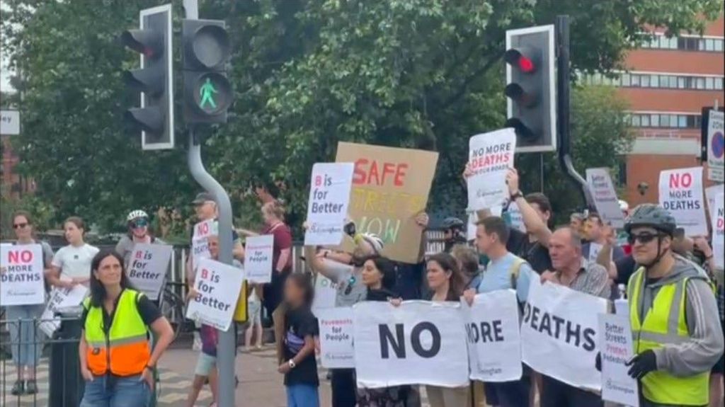 Dozens of members of the public stand on a road next to a pedestrian crossing whilst the light is red. they are holding up white signs that read "no more deaths" and "B is for Better"