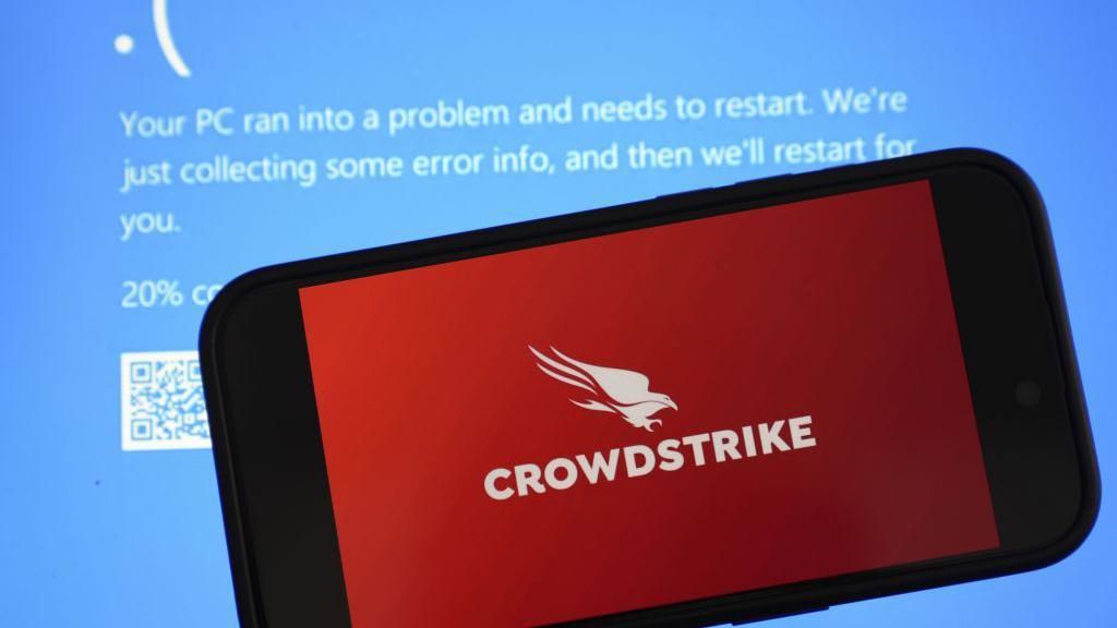 A phone with the red CrowdStrike logo of a bird of prey, placed in front of a PC screen displaying a blue error message
