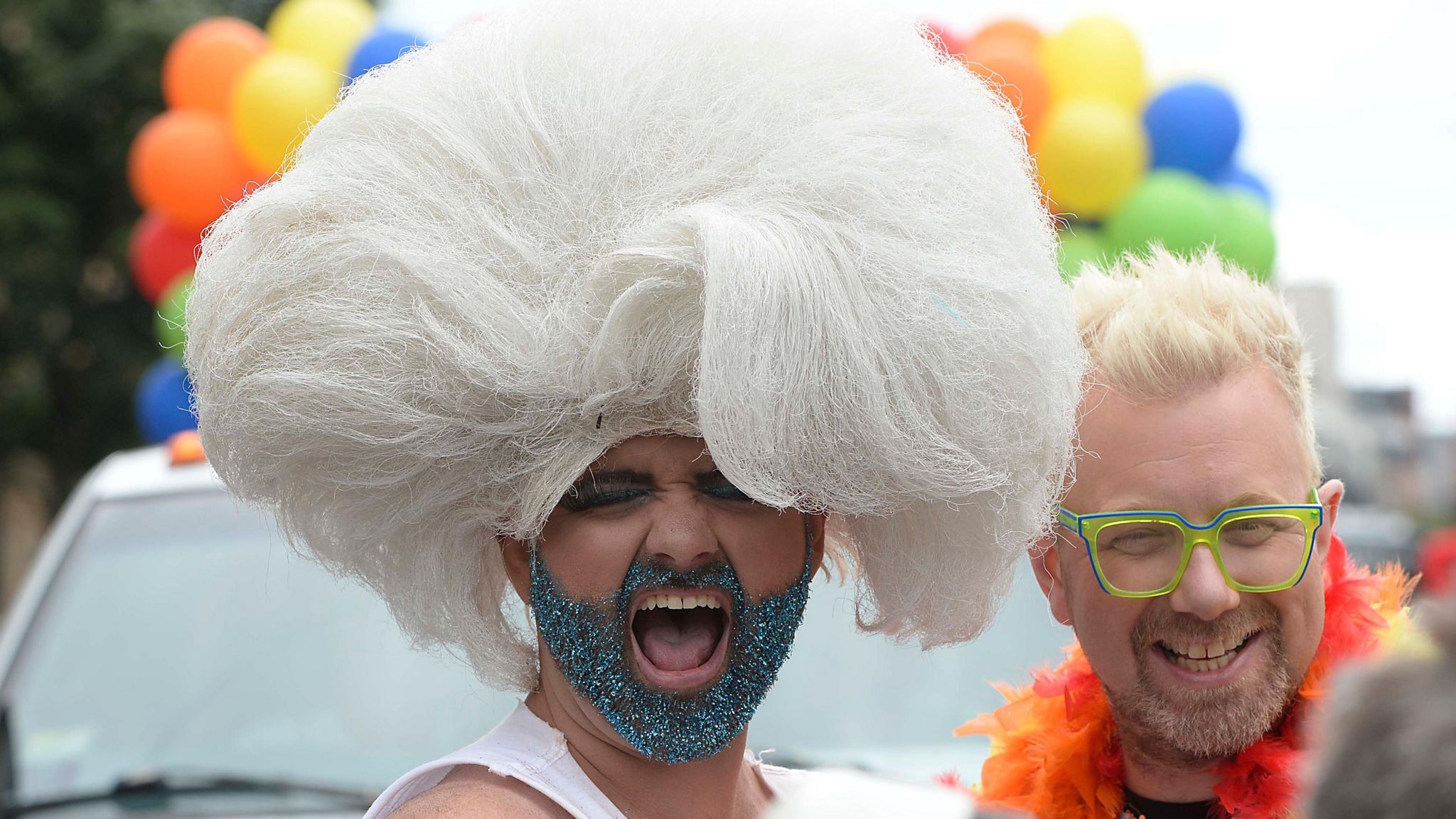 Two people in the parade. One has a white wig and a glittery beard. The other is wearing green glasses and wearing bright orange 