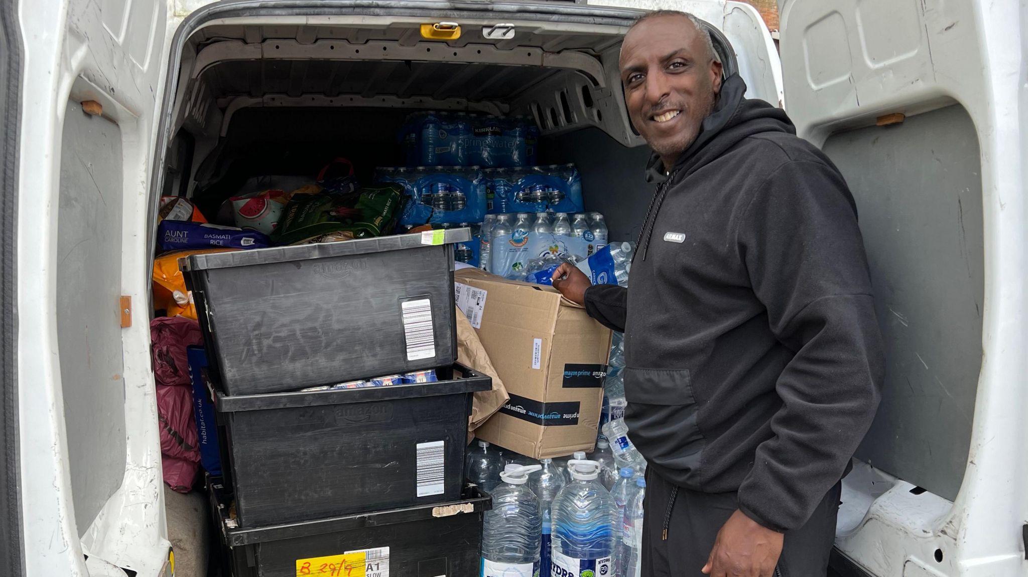 Orson De Silva, one of the aid organisers wearing a black jersey hoodie, with the supplies being gathered in Ladbroke Grove. He is standing by the back of a van full of boxes and water bottles