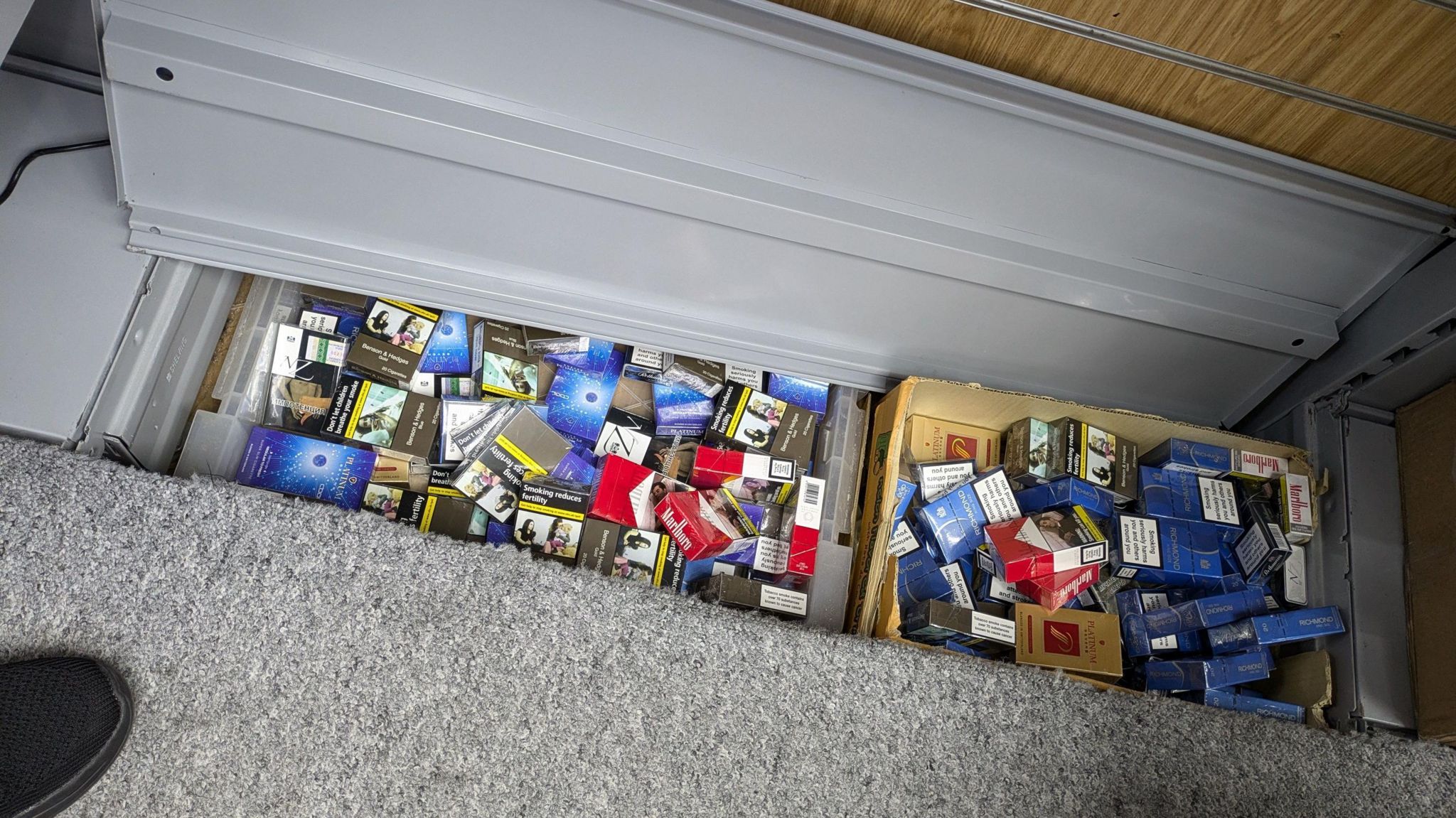 Two boxes stuffed with cigarettes in their packaging. The boxes are hidden under a grey floor panel which is lifted