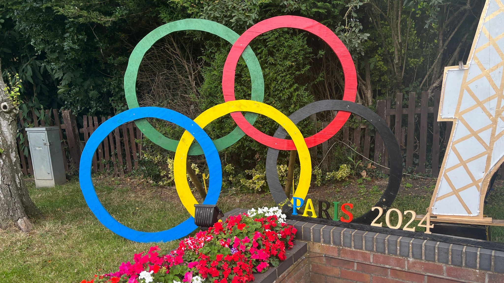 A model of the Olympic rings, sat behind a raised flower bed, with letters and numbers reading Paris 2024 on a wall