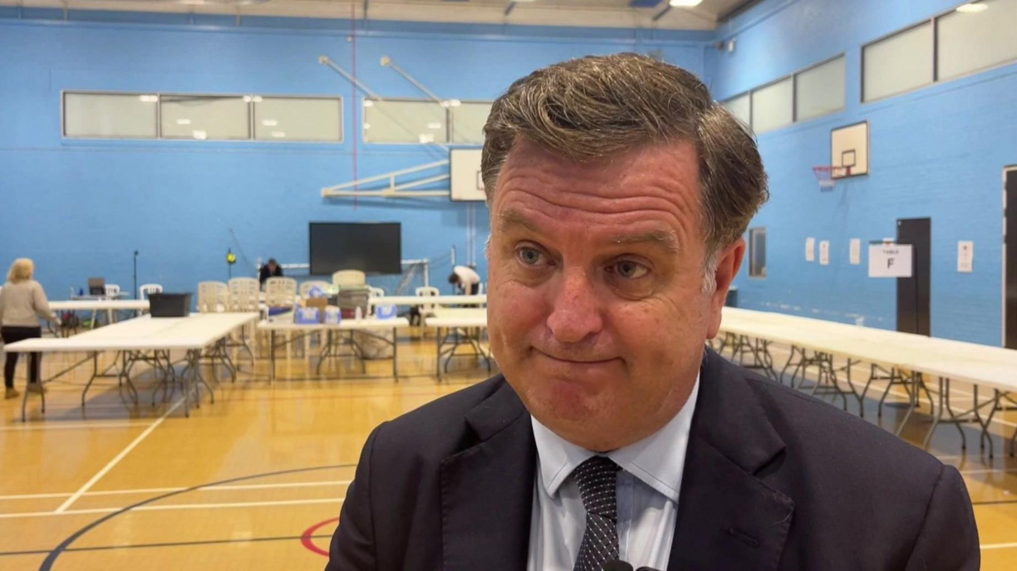 Close-up for Mel, a white man with grey hair with his mouth closed. He is in what appears to be a sports hall at a vote count. He looks disappointed. 