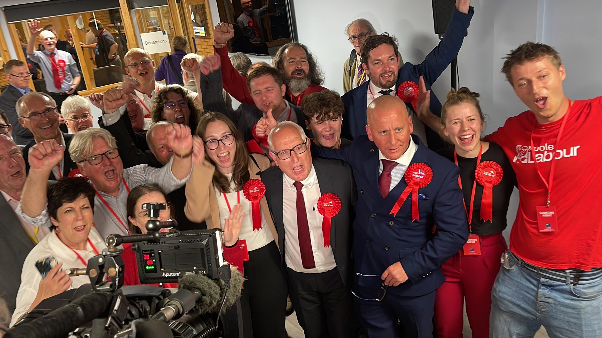 Room of people with red Labour rosettes cheering with camera in front of them.