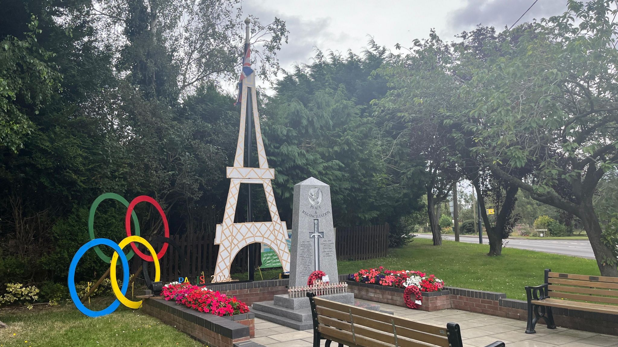A model of the Eiffel Tower and Olympic rings sit alongside a war memorial, with flower beds and benches in front