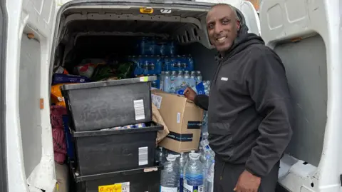 BBC Orson De Silva, one of the aid organisers wearing a black jersey hoodie, with the supplies being gathered in Ladbroke Grove. He is standing by the back of a van full of boxes and water bottles