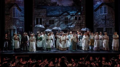 Set designed houses on the stage, they have brick walled features and crosses on their fronts and are designed to look Victorian. There are people on the stage with umbrellas and wearing victorian outfits. The orchestra can be seen at the front of the image.