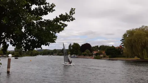 SATURDAY - A sailor in a white dinghy in the middle of the river at Cookham. There are trees overhanging the water on both sides of the bank and on the far side is a house with a red tile roof.