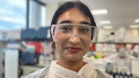 A teenage girl wearing laboratory goggles with a laboratory in soft focus behind her