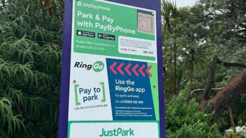 Blue, green and white parking sign with payment instructions - green trees and hedging behind