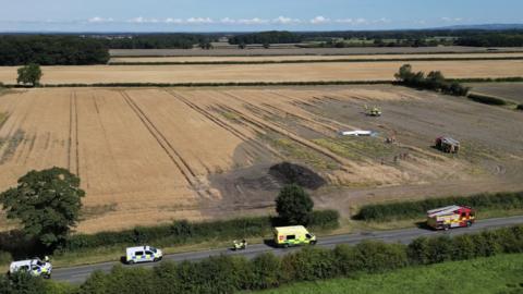 aerial image of plane crash in a field, a fire engine and air ambulance are in the field. On the road a number of emergency vehicles are in attendance. 