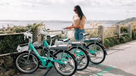 Three light green bikes standing up aside a path overlooking the coast, with a woman with sunglasses stood next to one, trying to get it started 