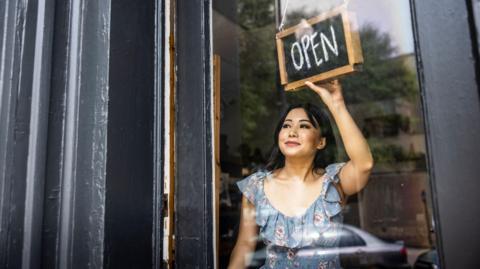 A woman wearing a blue floral top turns over a sign in her shop door saying Open