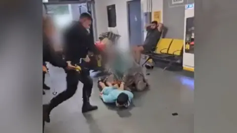 A police officer lifts his foot towards the head of a man who is lying on the floor
