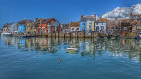 Jerry Rose MONDAY - Weymouth Harbour on the south coast is a typical English Harbour, here colourful buildings can be seen on the quay and are reflected in the water. In the foreground there is a small white rowing boat. The sky is blue and the sun is shining