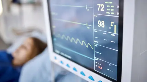 A heart monitor screen with a heartbeat rhythm. Out of focus, in the background, there is a woman laying on a hospital bed.