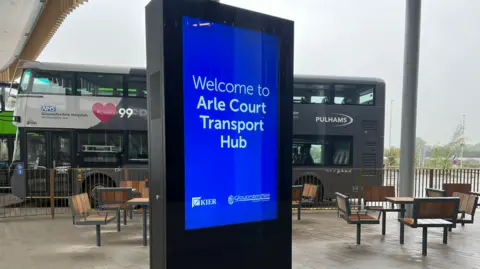 A sign displaying Arle Court Transport Hub with chairs, tables and a coach in the background