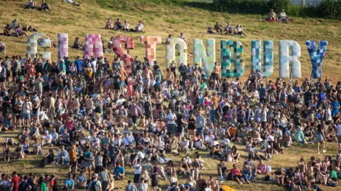 Getty Hundreds of people gathered, standing and sitting, on the grassy hill beneath the colourful Glastonbury sign. 