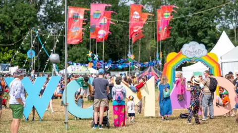 People walking past, climbing on and gathering around a colourful Womad sign
