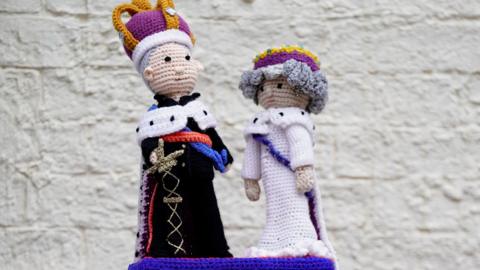 A woollen post box topper with figures of King Charles and Queen Camilla on top of a red post box