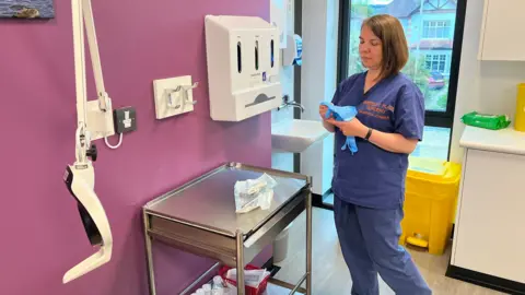 Dr Atkinson in blue scrubs standing near and sink and holding blue rubber gloves