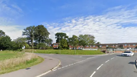 Junction of Heath House Lane and Barnfield Way, a T-junction on a bend with open land adjacent to the road and terraced housing in the background