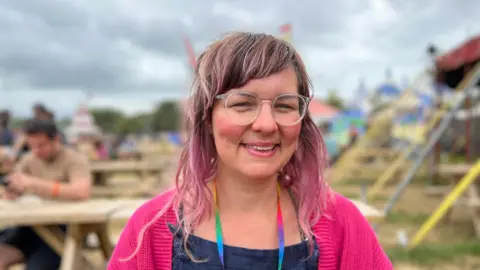 BBC Women with pink cardigan smiling, with tables and tent behind her.