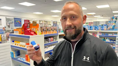 Bradley Belfit standing in a pharmacy holding an asthma inhaler, he is wearing a black zip-up jacket