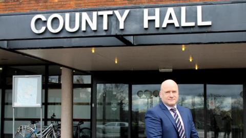 Man in navy blue suit standing outside building with the words "COUNTY HALL" In bold white lettering 