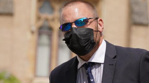 Craig Carter wearing a black face mask, blue tinted glasses and a navy jacket, striped shirt, navy tie walking into court on Friday 26 July