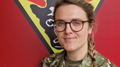 A female army officer with glasses and long plaited hair, wearing a camouflage army uniform