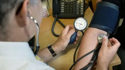 A doctor sitting at a desk taking a patient's blood pressure. He is holding a stethoscope against the patient's arm, which has a dark blue pad wrapped around the elbow area secured with Velcro. The doctor is holding a pressure gauge in his other hand.