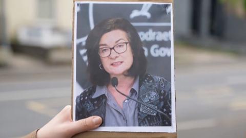 A hand holding a printed out picture of Eluned Morgan standing in front of a microphone