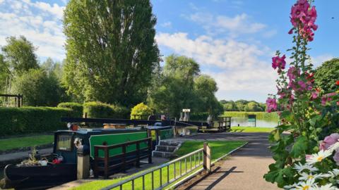 A green and black canal barge sits in Northmoor Lock with green hedges and trees behind it. The foreground sees tall pink hollyhock flowers and white daisies. The sky is sunny, blue and dappled white clouds.
