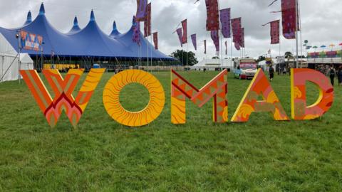 A colourful WOMAD sign in the middle of a field at a festival