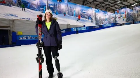 A girl with a prosthetic leg on a ski slope, holding her skiis