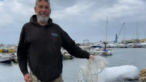Volunteer Dave Taylor on Poole harbourside with a handful of white gill netting with various boats on the water behind him