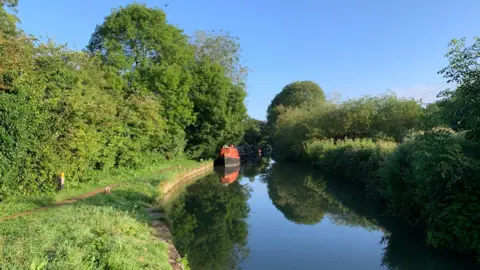 A sunny shot of the canal under a bright blue sky with a red narrowboat towards the horizon and a grassy footpath on the left.