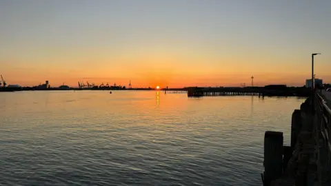 Tereza Rasova MONDAY - The sun sets over Southampton docks, on the horizon the sun appears as a small orange ball and the sky is glowing orange, you can see the silhouettes of cranes. In the foreground is the water in the harbour with the sun reflected in the ripples. There is a pier stretching across the middle of the picture from right to left.