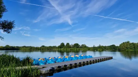 DereksDisco WEDNESDAY - A lake with a line of blue boats along a pontoon at Woodley. There are green plants in the water on the left of the frame and on the far side of the lake there are trees with green leaves against the blue sky.