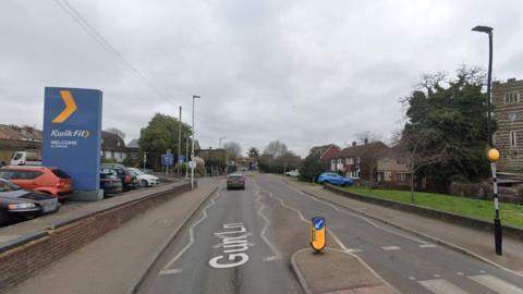A road. Foreground is a zebra crossing. To the left is a blue sign reading KwikFit. There is a church tower and houses on the right. The sky is grey.